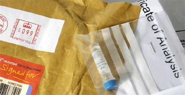 La imagen muestra un paquete abierto con un sobre de plástico transparente dentro, que parece contener un tubo de ensayo o muestrario médico con una tapa azul, insinuando la recepción de un componente o muestra para análisis.
