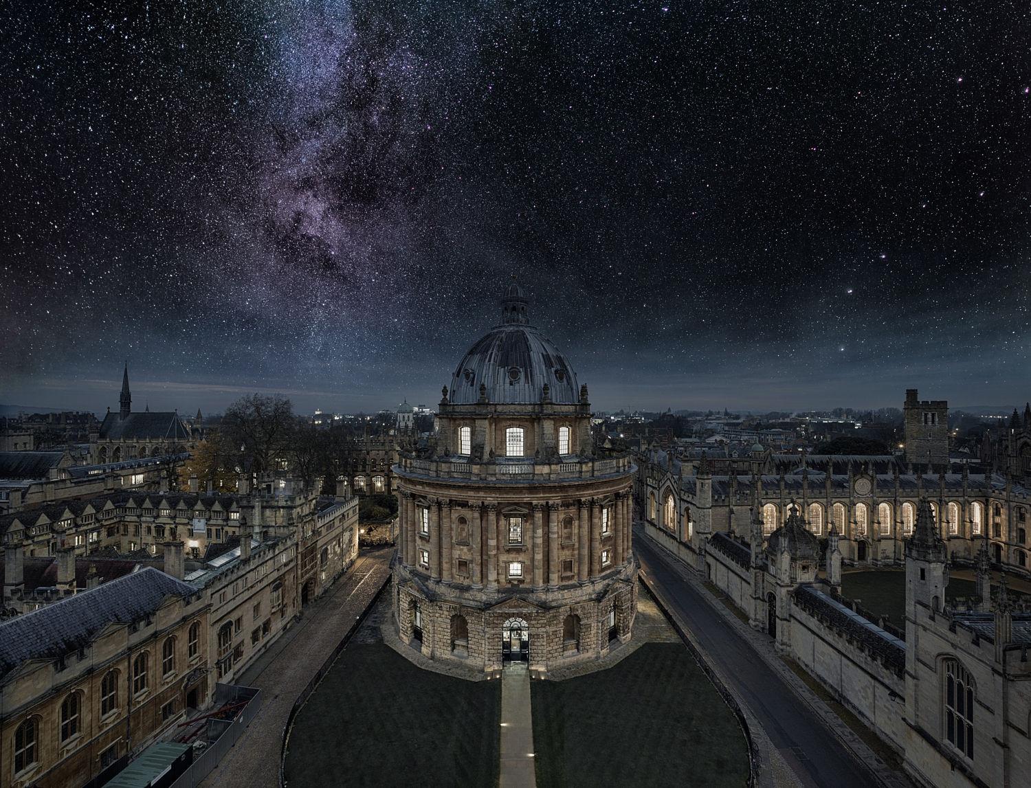 La imagen muestra una vista aérea nocturna de la Radcliffe Camera, parte de la Biblioteca Bodleiana de la Universidad de Oxford, con un cielo estrellado y la Vía Láctea visible en el fondo.