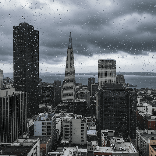 Vista del horizonte de San Francisco a través de una ventana cubierta de gotas de lluvia. La ciudad parece gris bajo un cielo nublado, con la bahía visible más allá de los edificios.