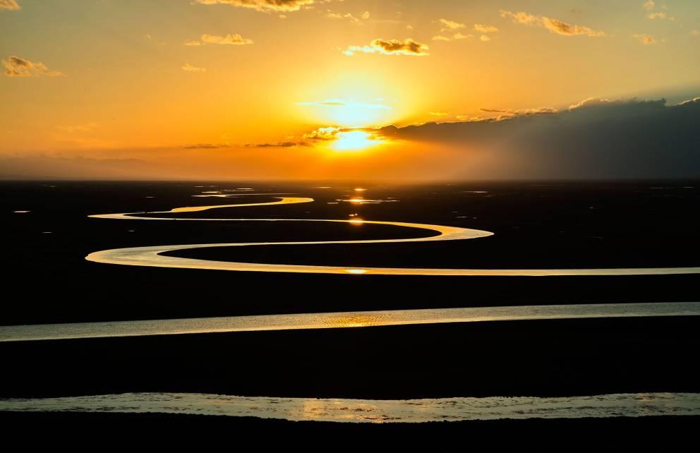 La imagen muestra un hermoso atardecer con el sol situado cerca del horizonte, proyectando rayos de luz a través de nubes dispersas. En primer plano, se observan varias curvas sinuosas de un río o humedal reflejando la luz dorada del sol.