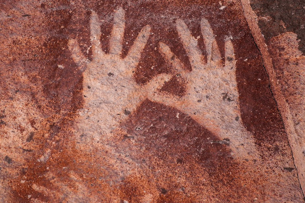 La imagen muestra dos impresiones de manos humanas en una superficie rocosa, que constituye una forma de arte rupestre en color rojo oscuro.