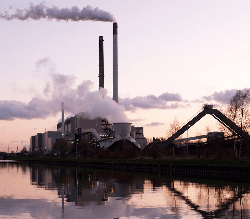 La imagen muestra una planta industrial con chimeneas emitiendo humo al atardecer, reflejándose sobre el agua de un río cercano.