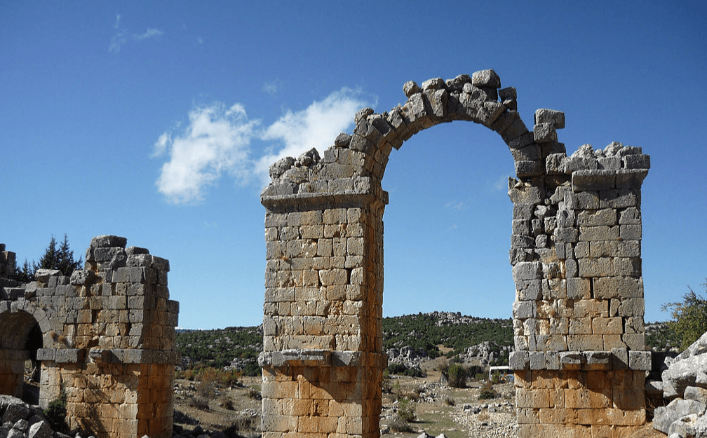 La imagen muestra las ruinas de un antiguo arco de piedra con un cielo azul parcialmente nublado de fondo. Las piedras del arco están erosionadas y el paisaje circundante es rocoso con vegetación baja.