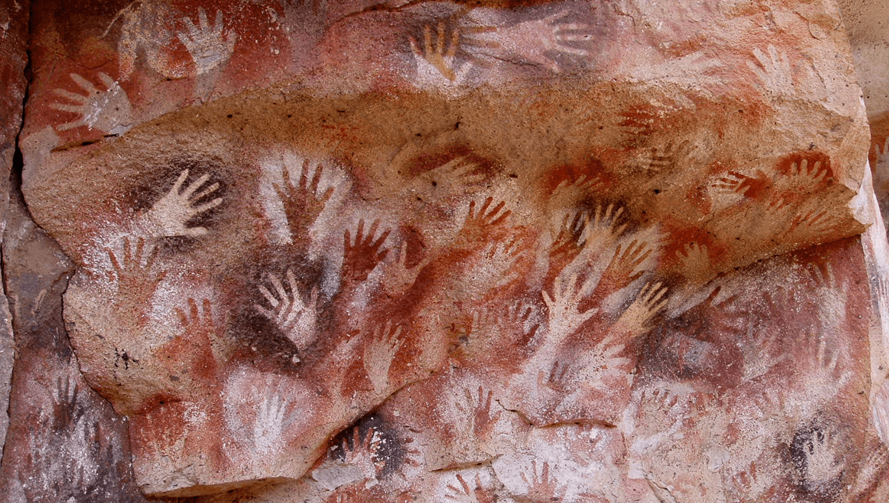 La imagen muestra una colección de impresiones de manos en una pared de roca, creadas mediante técnicas de pintura rupestre que dejan el contorno de las manos en colores blanco y rojo oscuro.