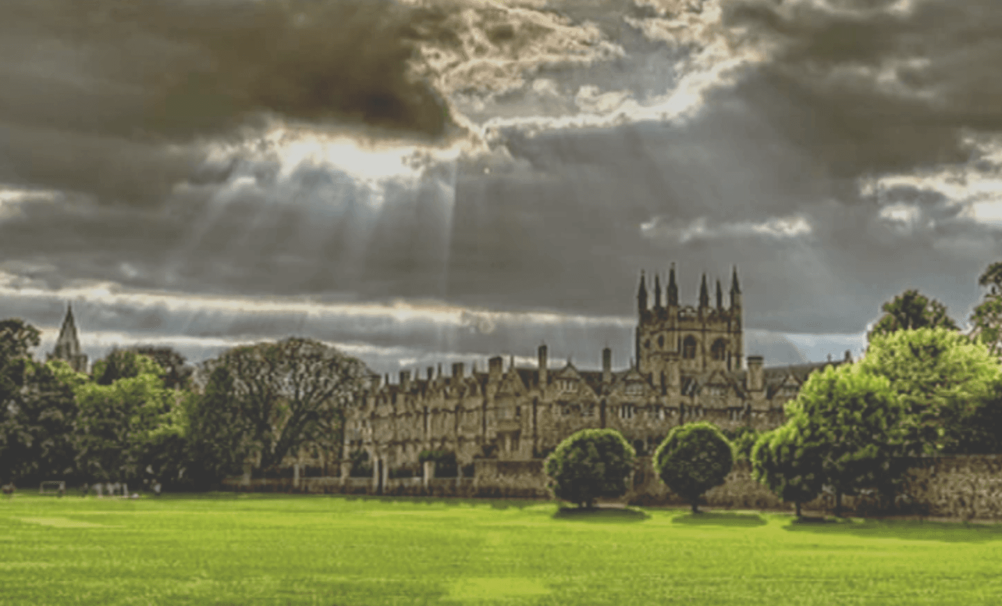 La imagen muestra un campo verde amplio con una edificación de estilo gótico al fondo, bajo un cielo nuboso por el cual se filtran rayos de sol.