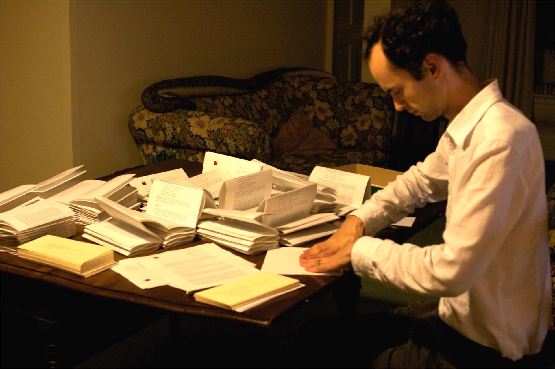 La imagen muestra a Toby Ord sentado frente a una mesa llena de papeles y documentos, ocupado en enviar formularios a la gente.