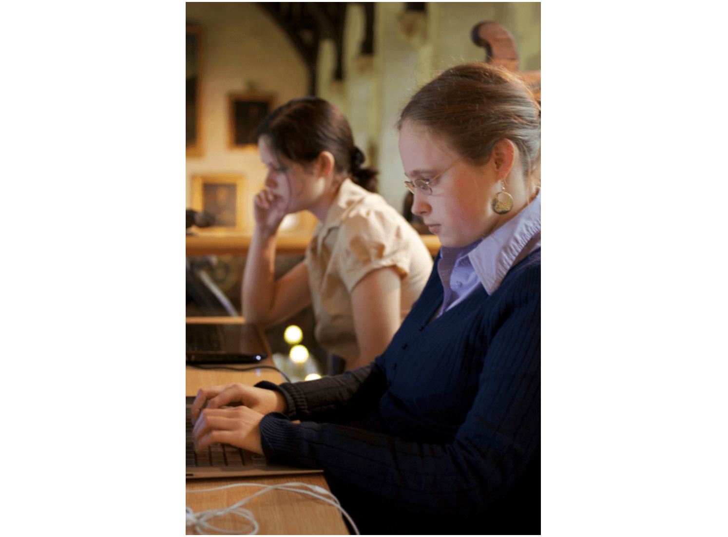 La imagen muestra a dos mujeres concentradas utilizando computadoras en un entorno que parece ser una oficina o biblioteca. La iluminación es tenue y la atmósfera parece tranquila.