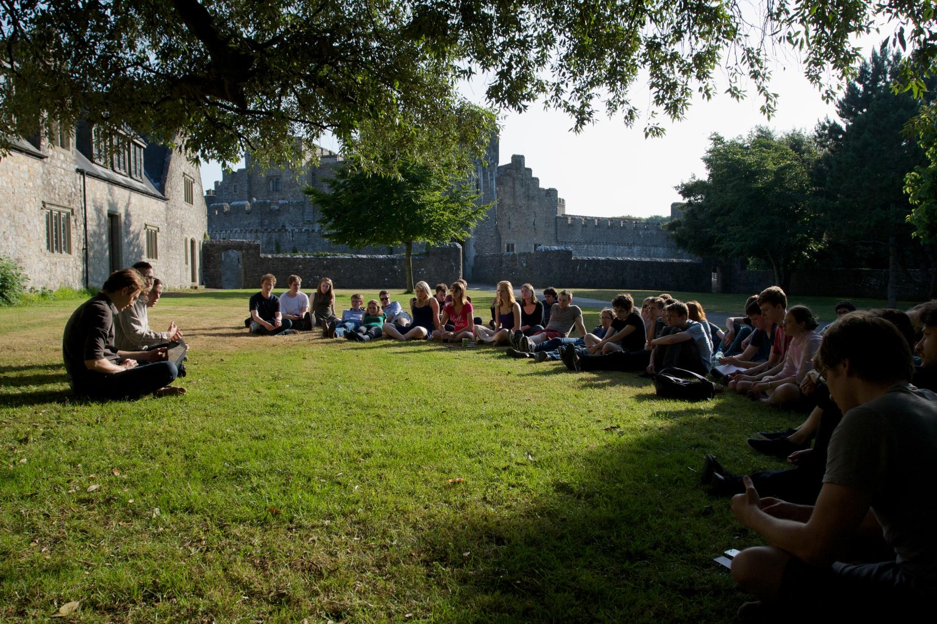 Un grupo de personas está sentado en círculo en un pasto verde, escuchando a un orador en un entorno que parece ser un campus o parque histórico con antiguos edificios de piedra al fondo.