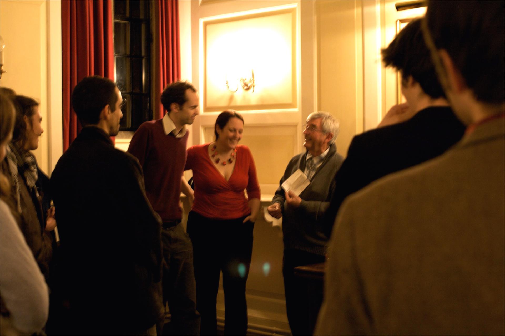 La imagen muestra un grupo de personas conversando en una sala con decoración clásica y una lámpara de pared encendida; se observa un ambiente social y cordial.