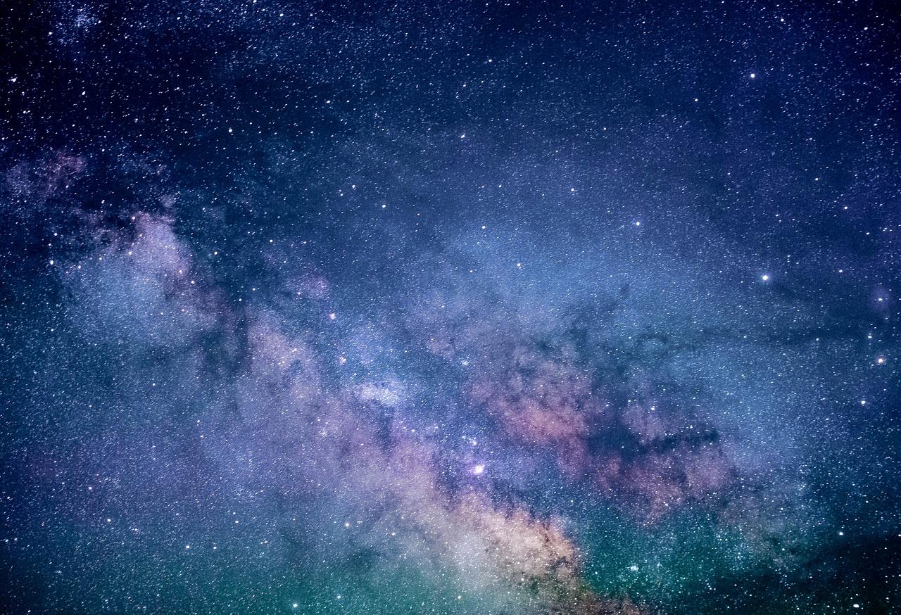 La imagen muestra una vista del cielo estrellado con la Vía Láctea claramente visible, presentando un mosaico de estrellas, nubes de gas y polvo interestelar en tonos de azul y púrpura.