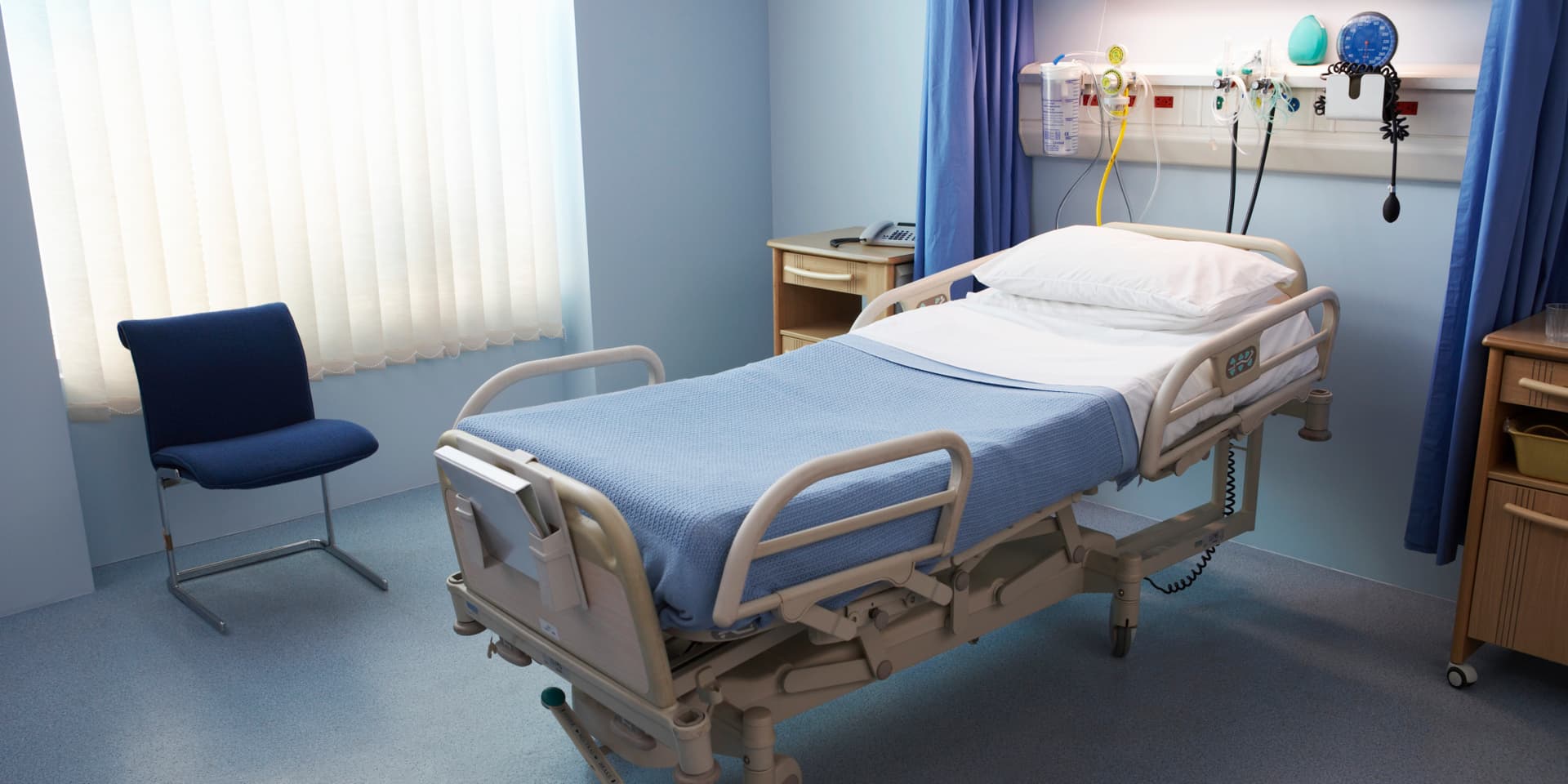 Una moderna habitación de hospital cuenta con una cama blanca ajustable con manta azul, una mesita de noche de madera con teléfono y una silla azul marino para visitantes cerca de las persianas verticales. El equipo médico, que incluye una bolsa de suero, máscaras de oxígeno y dispositivos de monitorización, está montado en la pared sobre la cama, separado por cortinas azules de privacidad.