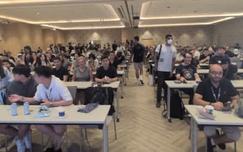 Una foto de una sala de conferencias abarrotada en Devcon con asistentes sentados en mesas y de pie junto a las paredes, viendo una charla.