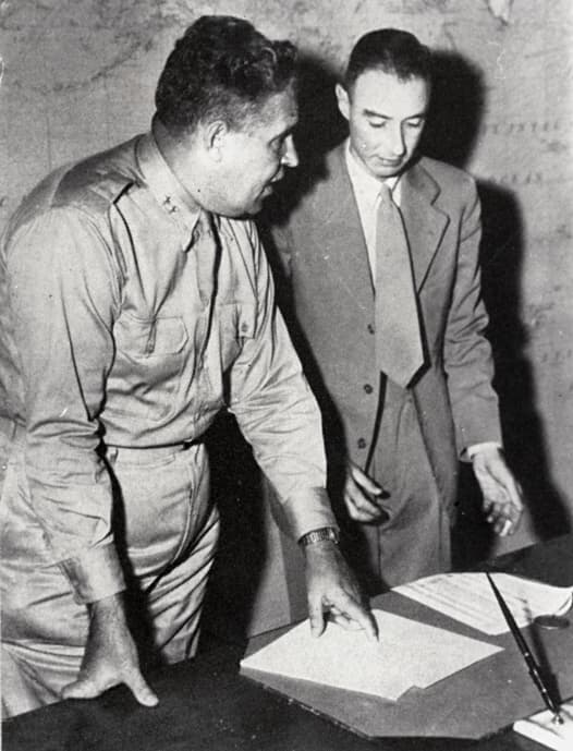 Fotografía en blanco y negro de dos hombres de pie junto a una mesa frente a un mapa mural. A la izquierda, un oficial militar uniformado, de pelo corto y oscuro, se inclina hacia delante y señala un documento que yace sobre la mesa. A la derecha, un hombre más delgado, con traje claro y corbata, mira el mismo documento mientras sostiene un bolígrafo.