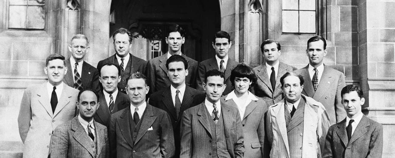 Fotografía de grupo en blanco y negro de unos diecisiete hombres y una mujer posando en las escaleras de un edificio de piedra de estilo gótico. Están dispuestos en tres filas y visten traje y corbata, en su mayoría de tonos claros y medios, con un arco de piedra tallada visible detrás de ellos.