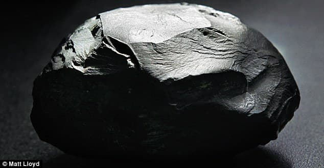 Fotografía de una roca redondeada y completamente negra sobre una superficie oscura, iluminada desde arriba de modo que sus facetas aparecen como reflejos grises pálidos, casi blancos, sobre sombras profundamente negras, lo que produce una impresión plana, tipo collage, de manchas blancas, grises y negras. Texto: Matt Lloyd.