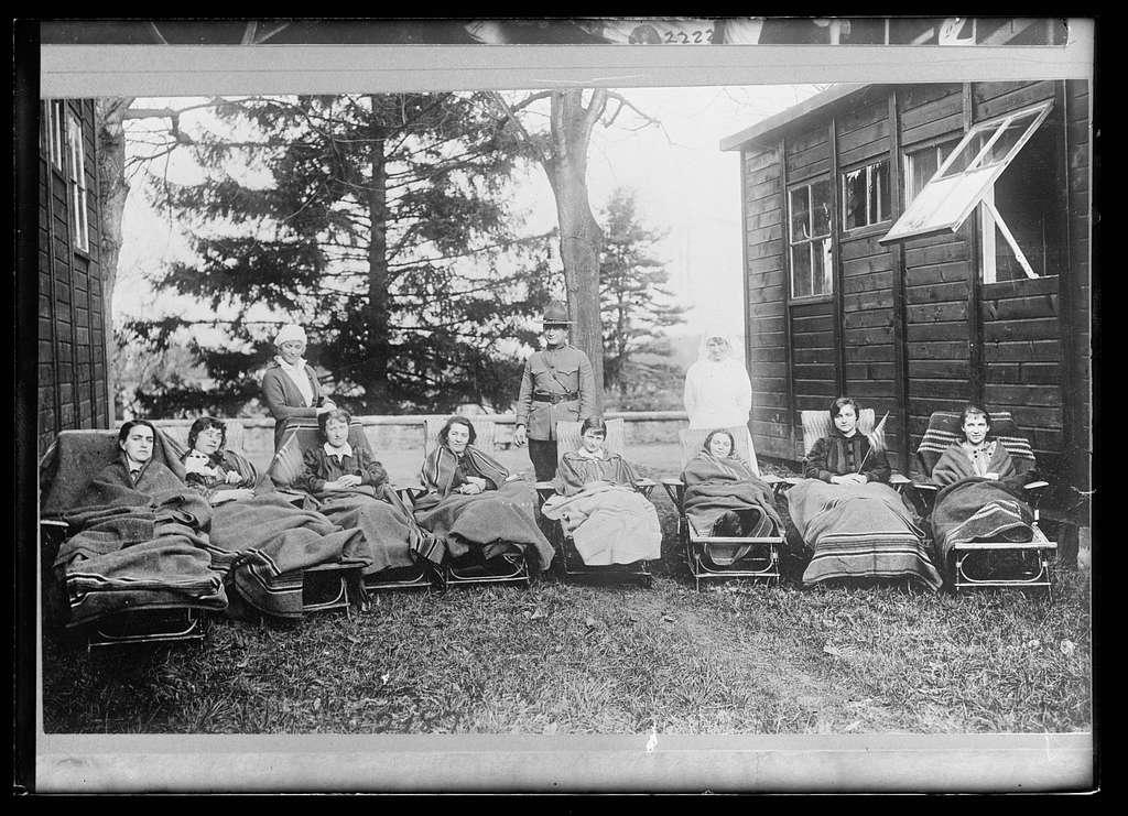 En una fotografía histórica en blanco y negro, un grupo de varias personas está reunido al aire libre. Un hombre con uniforme militar está de pie en el centro, flanqueado por dos mujeres vestidas de blanco, probablemente trabajadoras sanitarias. Las otras mujeres están sentadas en tumbonas, envueltas en mantas, lo que sugiere un momento de descanso o recuperación. Al fondo, hay árboles altos y una estructura de madera, probablemente un edificio o refugio, con ventanas. La escena transmite una sensación de comunidad y cuidado, posiblemente en un contexto relacionado con la salud o la recuperación durante una época pasada. La atmósfera general es tranquila y de apoyo, ambientada en un entorno natural.