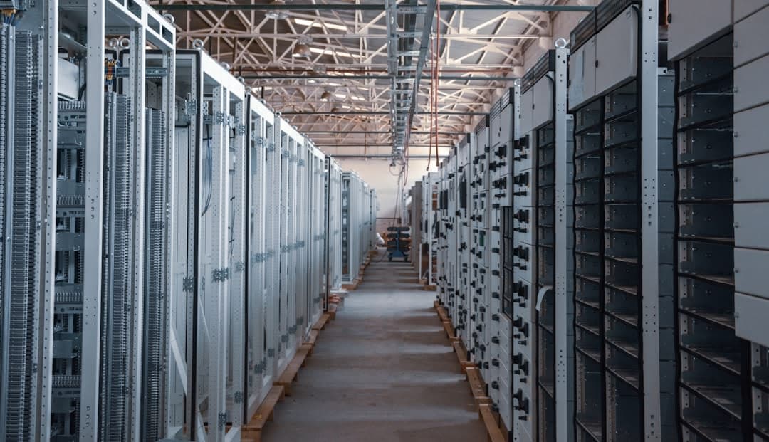 La imagen muestra un largo pasillo en lo que parece ser un centro de datos o una instalación eléctrica, con filas de racks de servidores y paneles de equipos eléctricos a ambos lados.