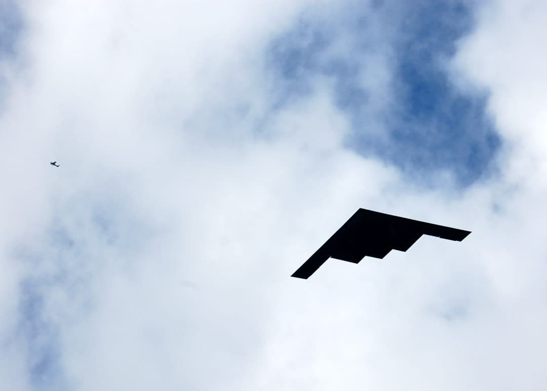 Un bombardero furtivo B-2 Spirit vuela contra un cielo nublado; su característico diseño de ala volante crea una nítida silueta negra. Se aprecia un pequeño pájaro en la esquina superior izquierda del encuadre, proporcionando una escala para el enorme avión.