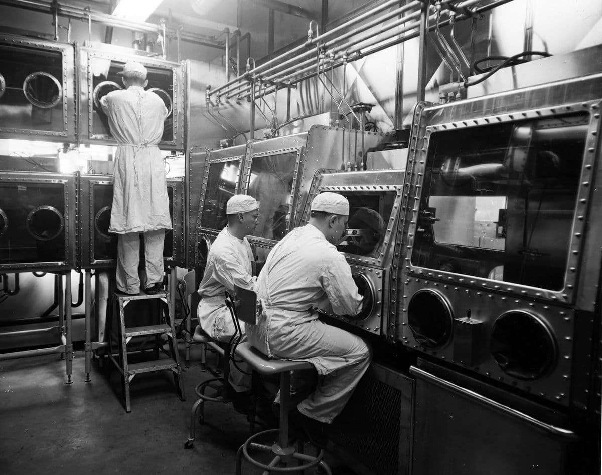 La imagen muestra a tres personas trabajando en un laboratorio con equipos de contención, todos visten ropa protectora y están manipulando material a través de compartimientos sellados con guantes incorporados.