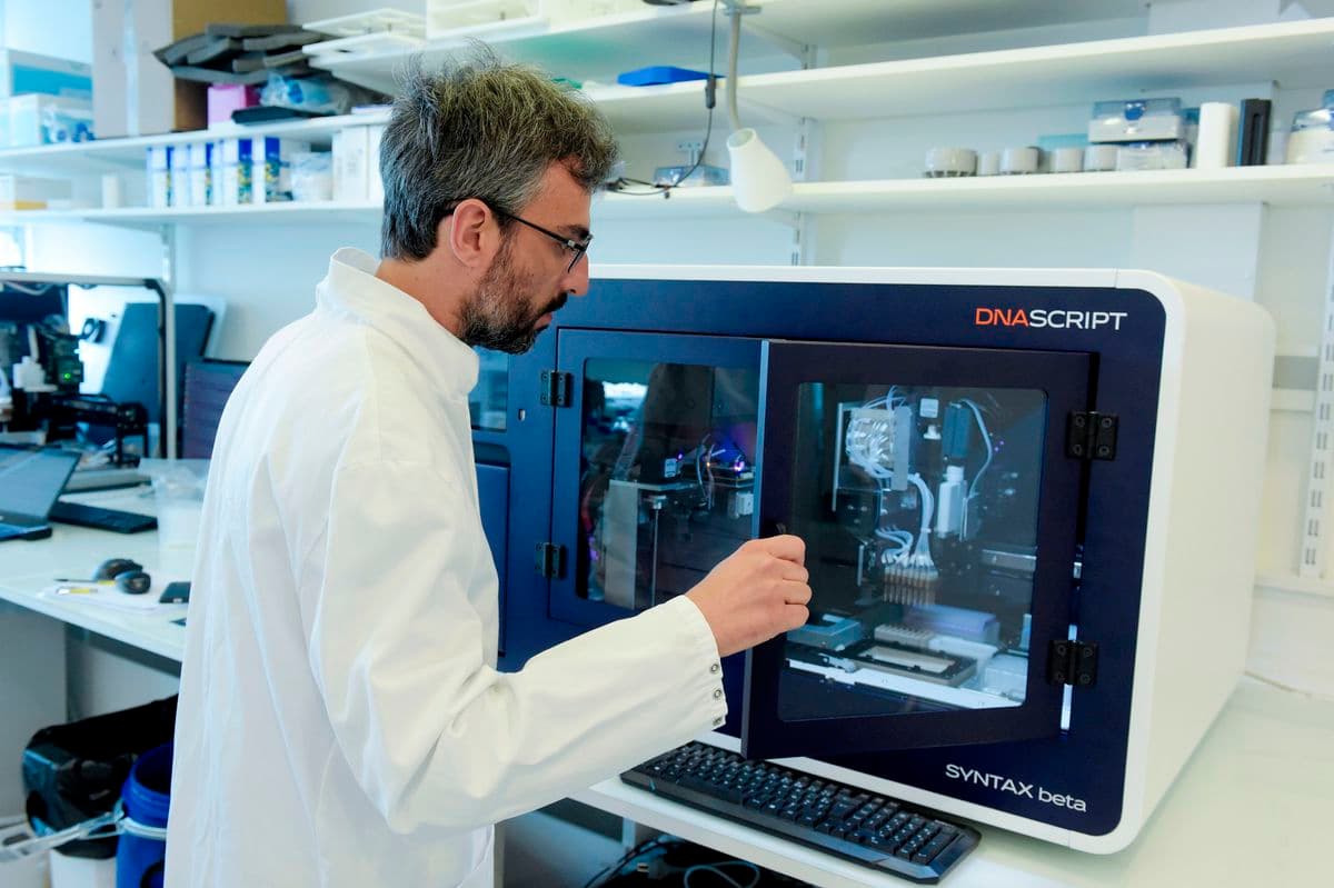Un hombre con bata de laboratorio está manipulando un equipo de laboratorio. Se encuentra en un laboratorio con estanterías llenas de suministros y equipo científico de fondo.