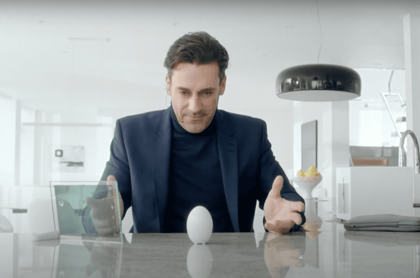 Un hombre vestido de traje está inclinado sobre una superficie pulida en una cocina moderna, observando con interés un aparato con forma de huevo colocado en forma horizontal. La escena sugiere una atmósfera de curiosidad o sorpresa.