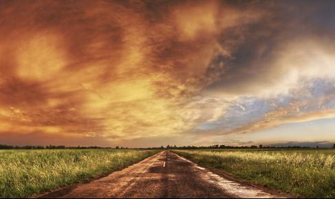 La imagen muestra un camino recto que atraviesa una planicie, flanqueado por campos verdes bajo un cielo que a la izquierda aparece dramático, con nubes que reflejan tonos anaranjados y amarillos, y a la derecha aparece claro y luminoso.