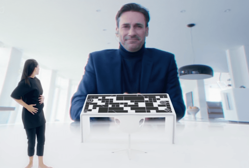 La imagen muestra una mujer que se encuentra frente a una pantalla de gran tamaño, en la que un hombre sonríe y se inclina hacia ella. Frente a la mujer hay una mesa de control de estilo futurista.