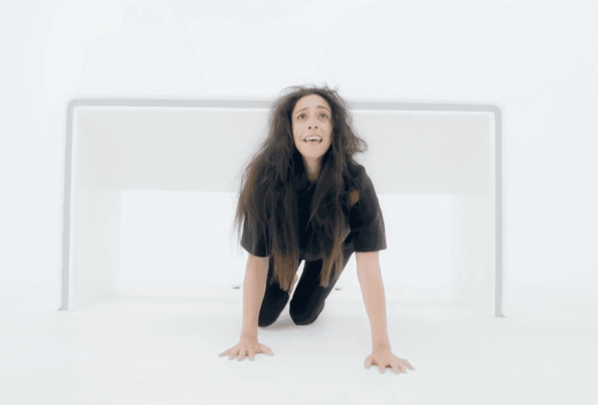 La imagen muestra a una mujer arrodillada y con las manos en el suelo en una habitación blanca, con una expresión que indica temor y sufrimiento.