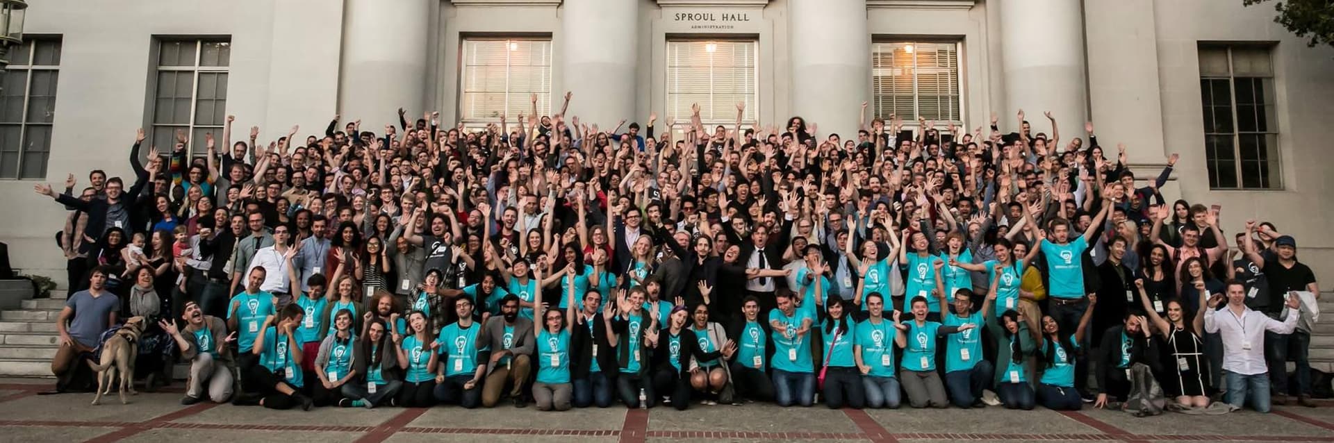 Un gran grupo de personas, muchas de ellas con camisetas turquesas a juego, posan con entusiasmo con los brazos en alto en las escaleras del Sproul Hall, un edificio con columnas clásicas. La multitud parece estar celebrando lo que parece ser un evento universitario o institucional.