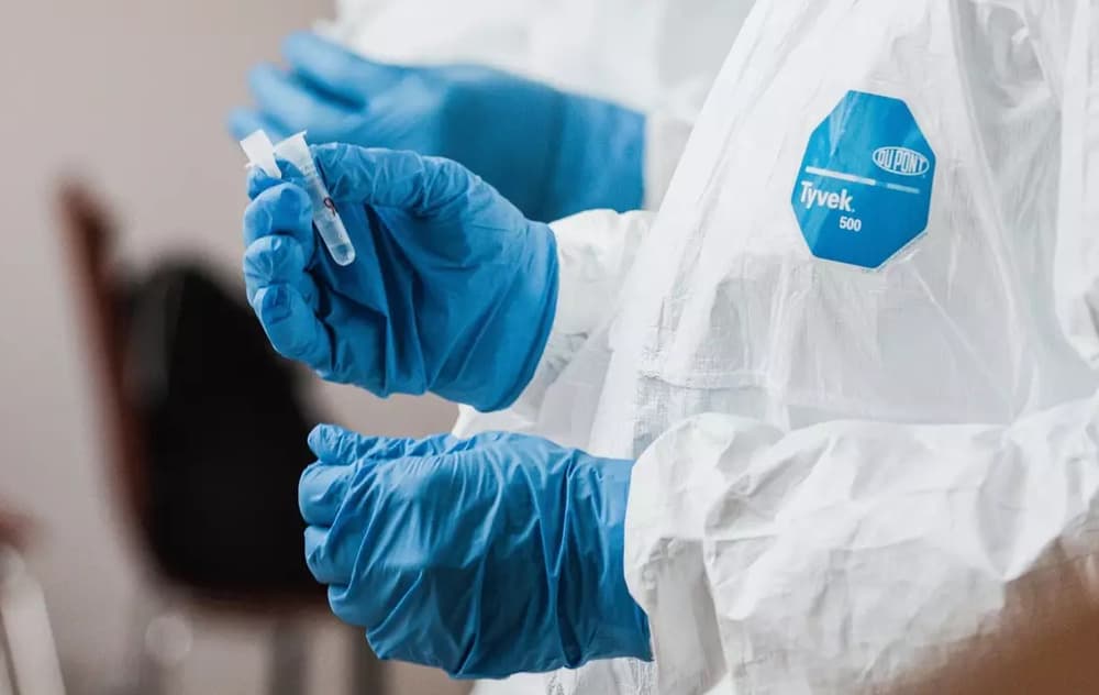 Un trabajador sanitario con un traje protector blanco Tyvek 500 y guantes azules de nitrilo sostiene un pequeño frasco médico o tubo de ensayo. La imagen captura el entorno clínico y estéril de las pruebas médicas o el trabajo farmacéutico, con la marca DuPont visible en el equipo de protección.