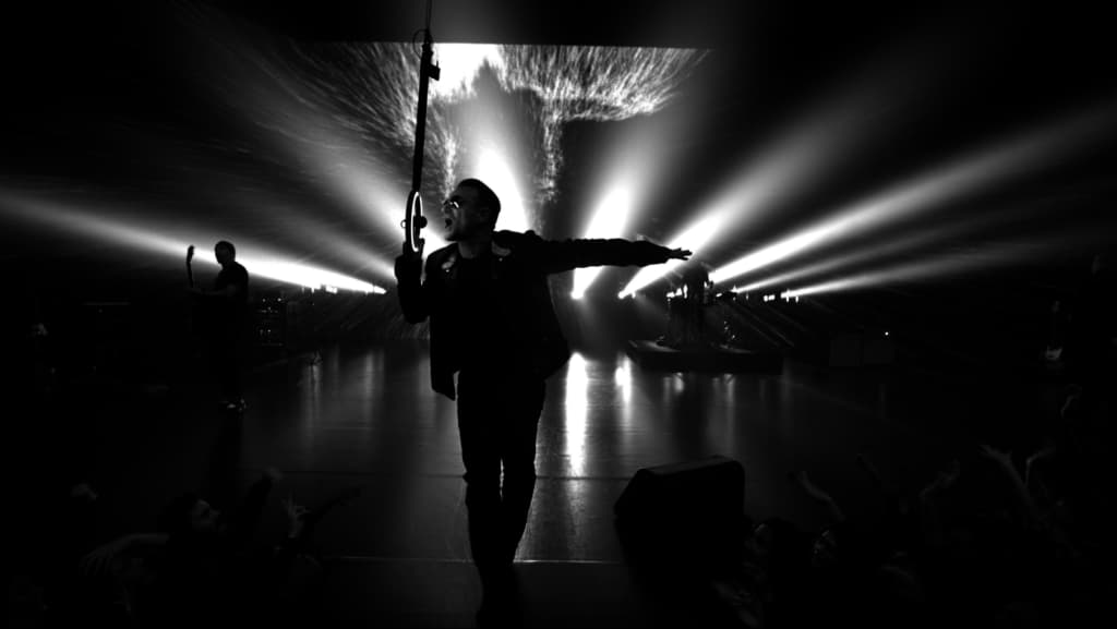Una dramática fotografía en blanco y negro de un concierto captura la silueta de un artista, de pie en el centro del escenario con los brazos extendidos, con un explosivo telón de fondo de luces escénicas. Potentes haces de luz irradian hacia afuera, mientras que una elaborada iluminación crea un efecto similar al de una fuente sobre su cabeza, creando una atmósfera teatral impactante.