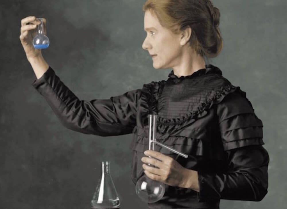Una mujer con un vestido negro de la época victoriana sostiene un frasco que contiene un líquido azul mientras lo examina, y sobre la mesa que tiene delante puede verse más material de vidrio de laboratorio. La imagen evoca el período histórico de los inicios de la investigación científica, presentada en un estilo de retrato sobre un fondo apagado.