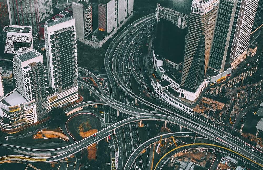 Vista aérea de un complejo intercambiador de autopistas con múltiples carriles curvos que se entrelazan entre modernos rascacielos en un entorno urbano. La imagen captura la intrincada infraestructura del transporte urbano con sus amplios pasos elevados y carreteras, con la arquitectura contemporánea como telón de fondo.