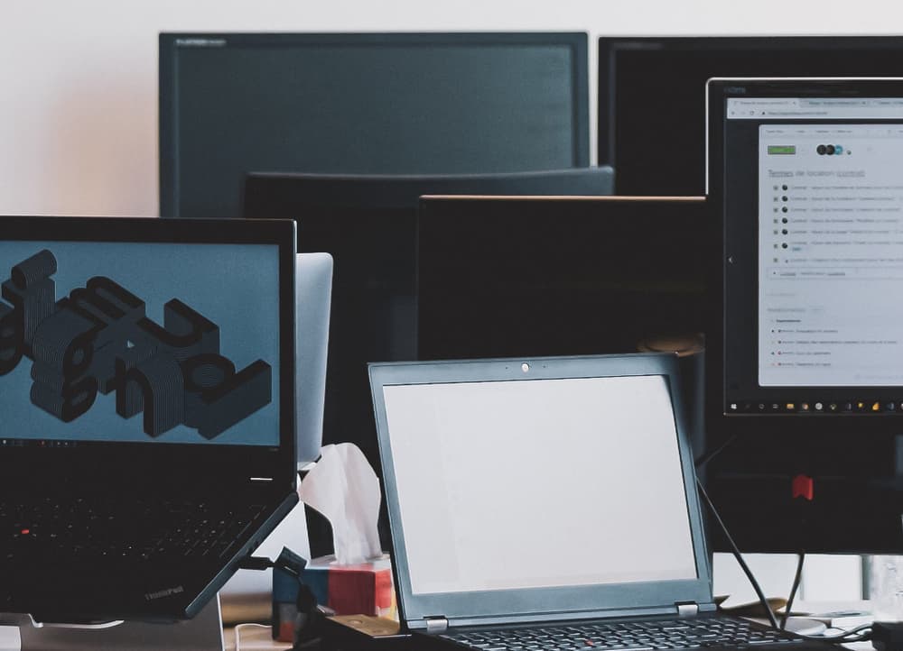 La imagen muestra un espacio de trabajo con múltiples monitores, ordenadores y pantallas. En primer plano hay un ordenador portátil con la pantalla en blanco, rodeado de otros monitores que muestran diversos contenidos, incluidos gráficos en 3D y lo que parece ser una página de documentación o un foro. La configuración sugiere un entorno de desarrollo profesional o de diseño.