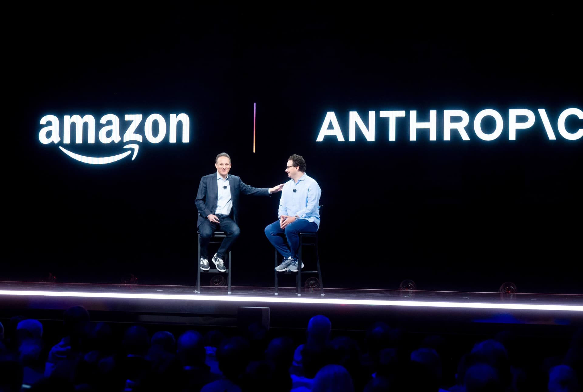 Dos hombres sentados en taburetes conversan en un escenario frente al público, bajo los logotipos de Amazon y Anthropic proyectados en una gran pantalla.